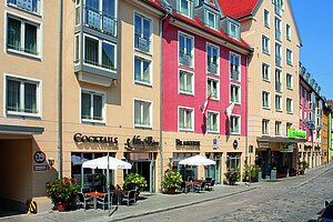 Farbiges Gebäudeensemble des Holiday Inn am Jakobsmarkt mit Glasbalkonen, Außengastronomie und gepflastertem Straßenbereich.