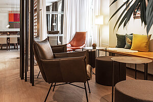 Moderner Lounge‑Bereich im Hotel am Jakobsmarkt mit Ledersesseln, rotem Designstuhl, bunten Kissen und hellen Fensterfronten.