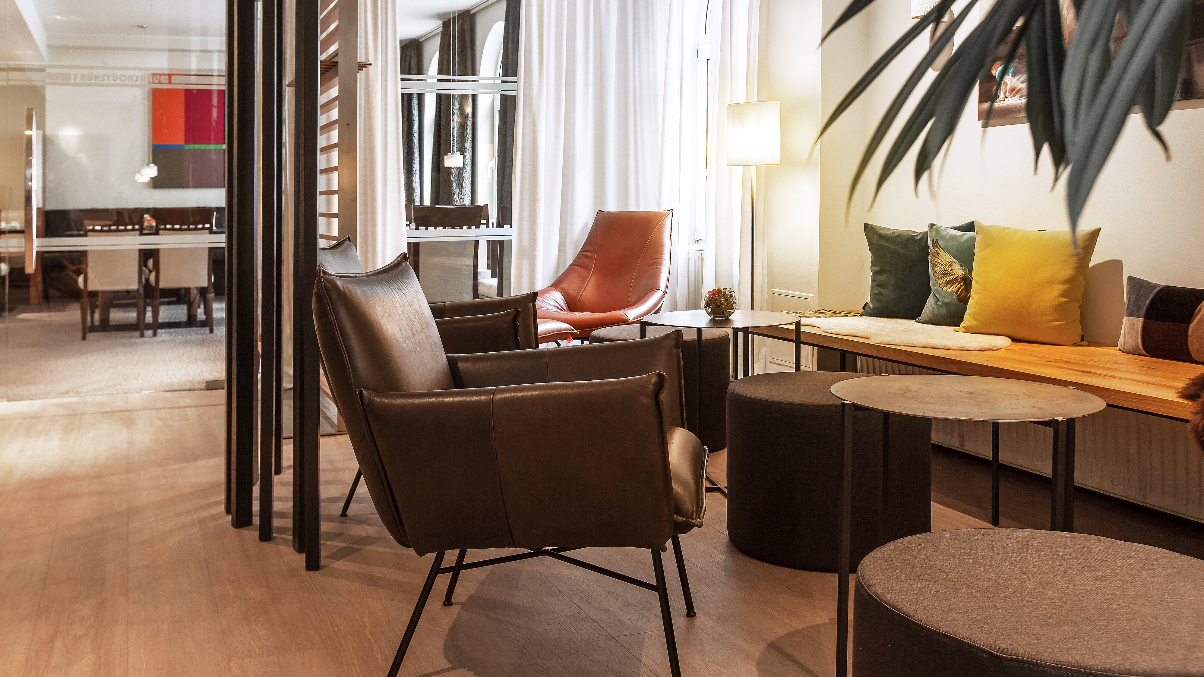 Moderner Lounge‑Bereich im Hotel am Jakobsmarkt mit Ledersesseln, rotem Designstuhl, bunten Kissen und hellen Fensterfronten.