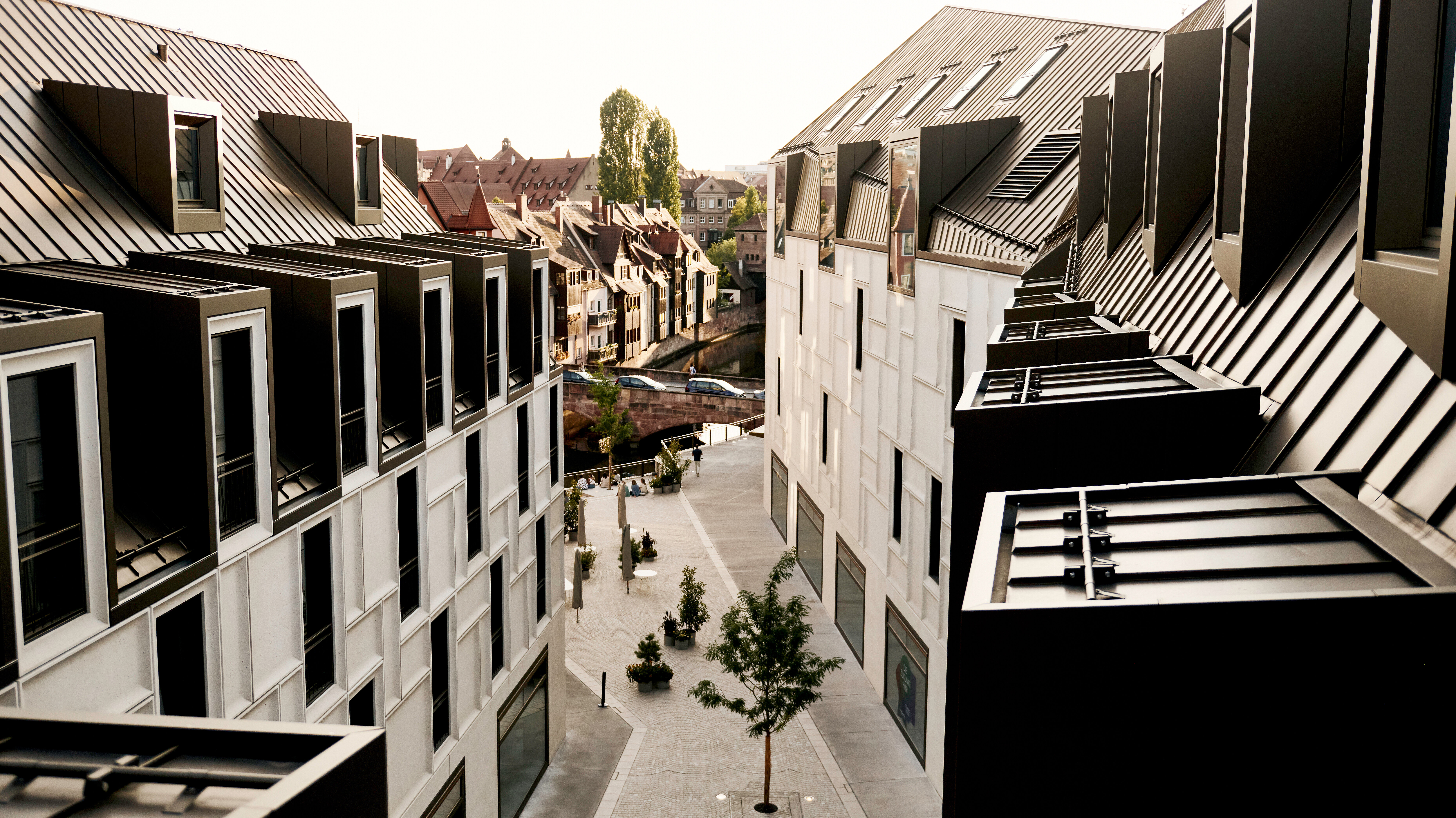 Blick aus der Höhe auf eine moderne Gasse im Augustinerhof mit weißen Fassaden und dunklen Metalldächern, die auf eine Brücke und historische Gebäude im Hintergrund zuführt.