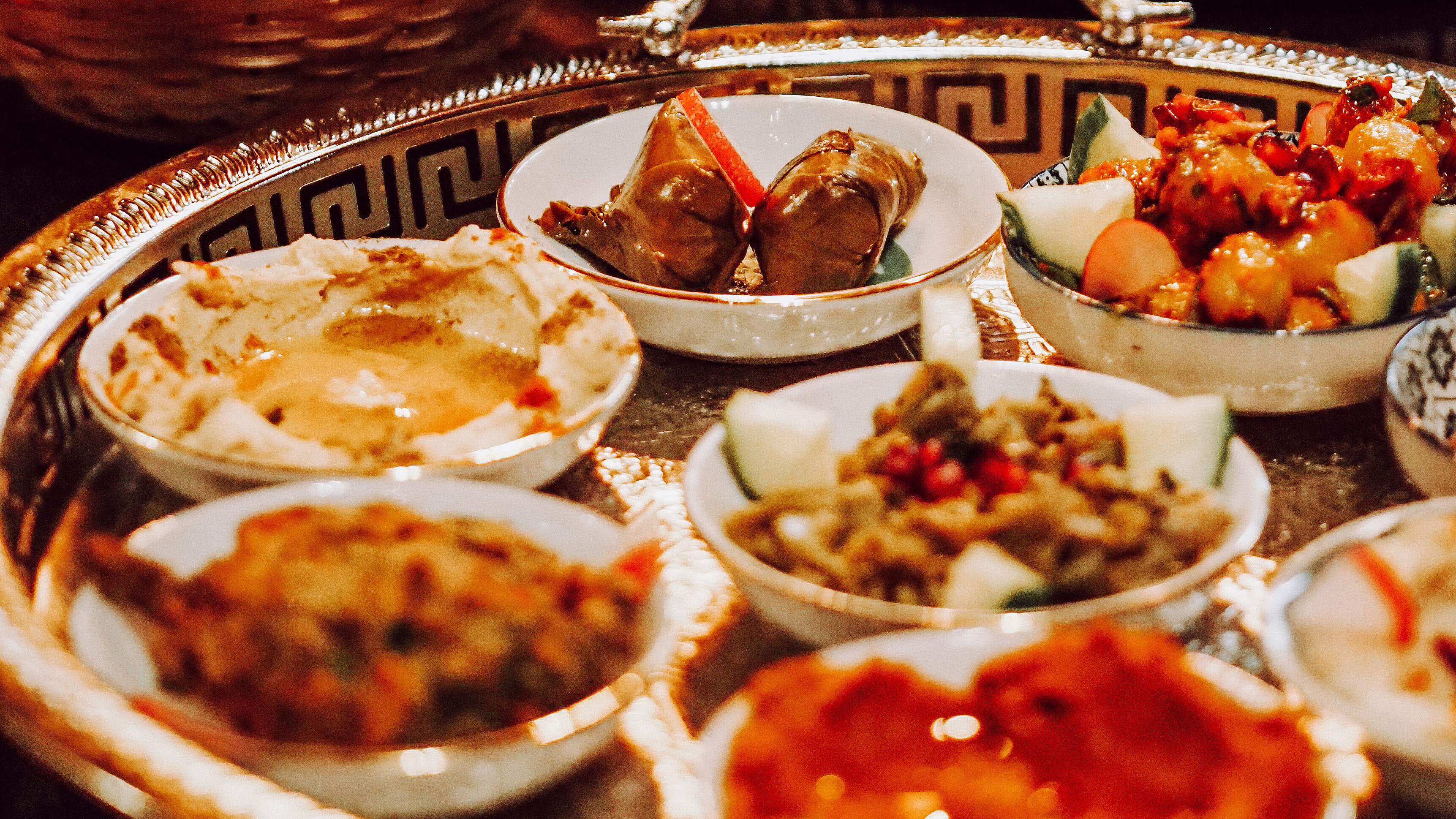 Ein reich gedeckter Tisch mit silbernen Serviertabletts voller levantinischer Mezze – darunter cremige Aufstriche, gefüllte Weinblätter, Salate und frittiertes Gebäck – daneben ein Korb mit Fladenbrot und frischem Gemüse.