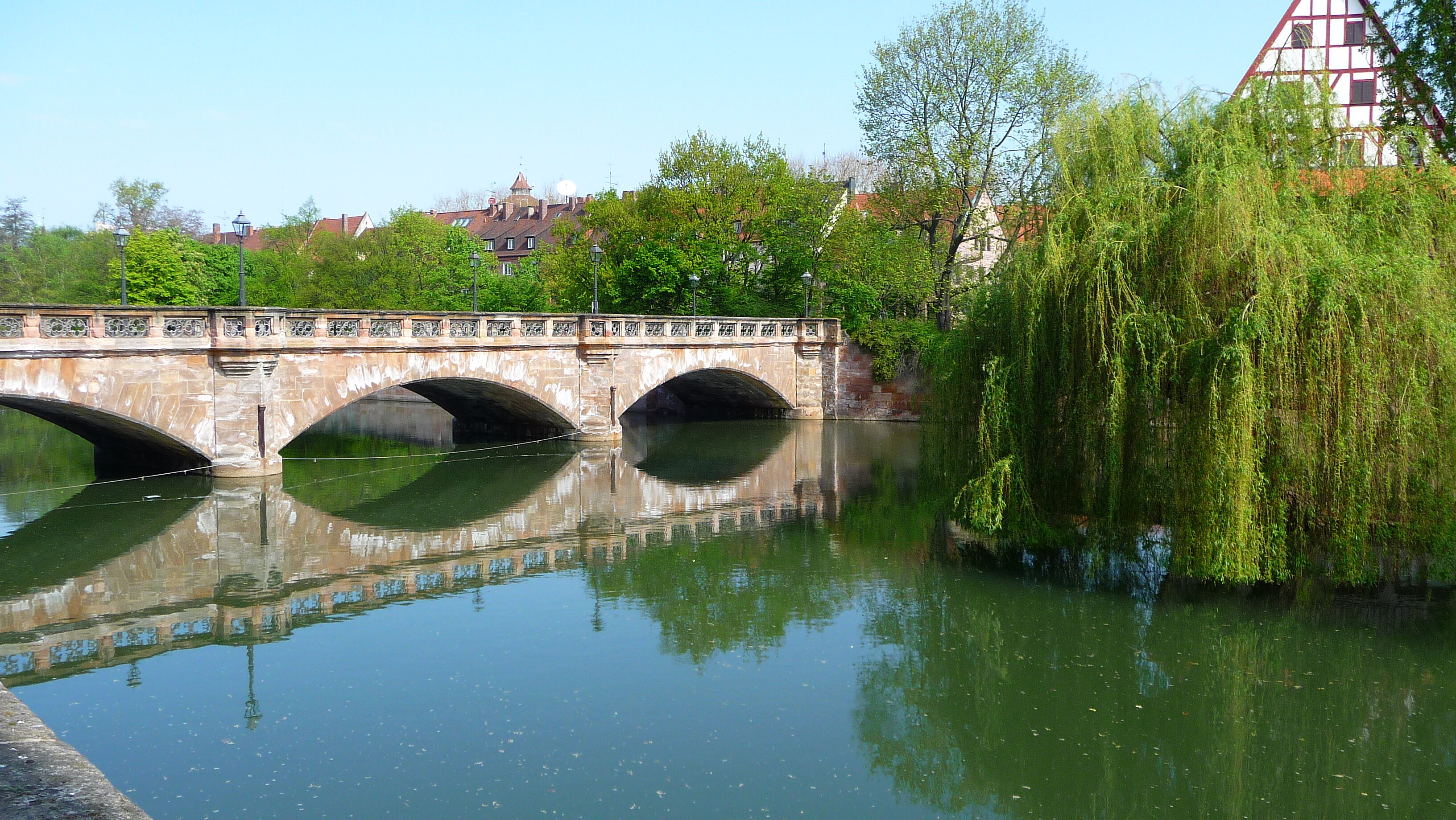 Nürnberger Maxbrücke: Die Sandsteinbogenbrücke spiegelt sich an einem sonnigen Frühlingstag in der Pegnitz, flankiert von einer grünen Trauerweide.