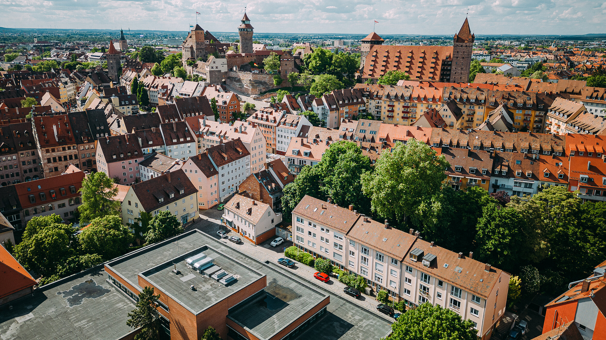 Das Luftbild zeigt die Nürnberger Kaiserburg und die umliegende Altstadt mit ihren markanten roten Ziegeldächern unter einem sonnigen, leicht bewölkten Himmel.