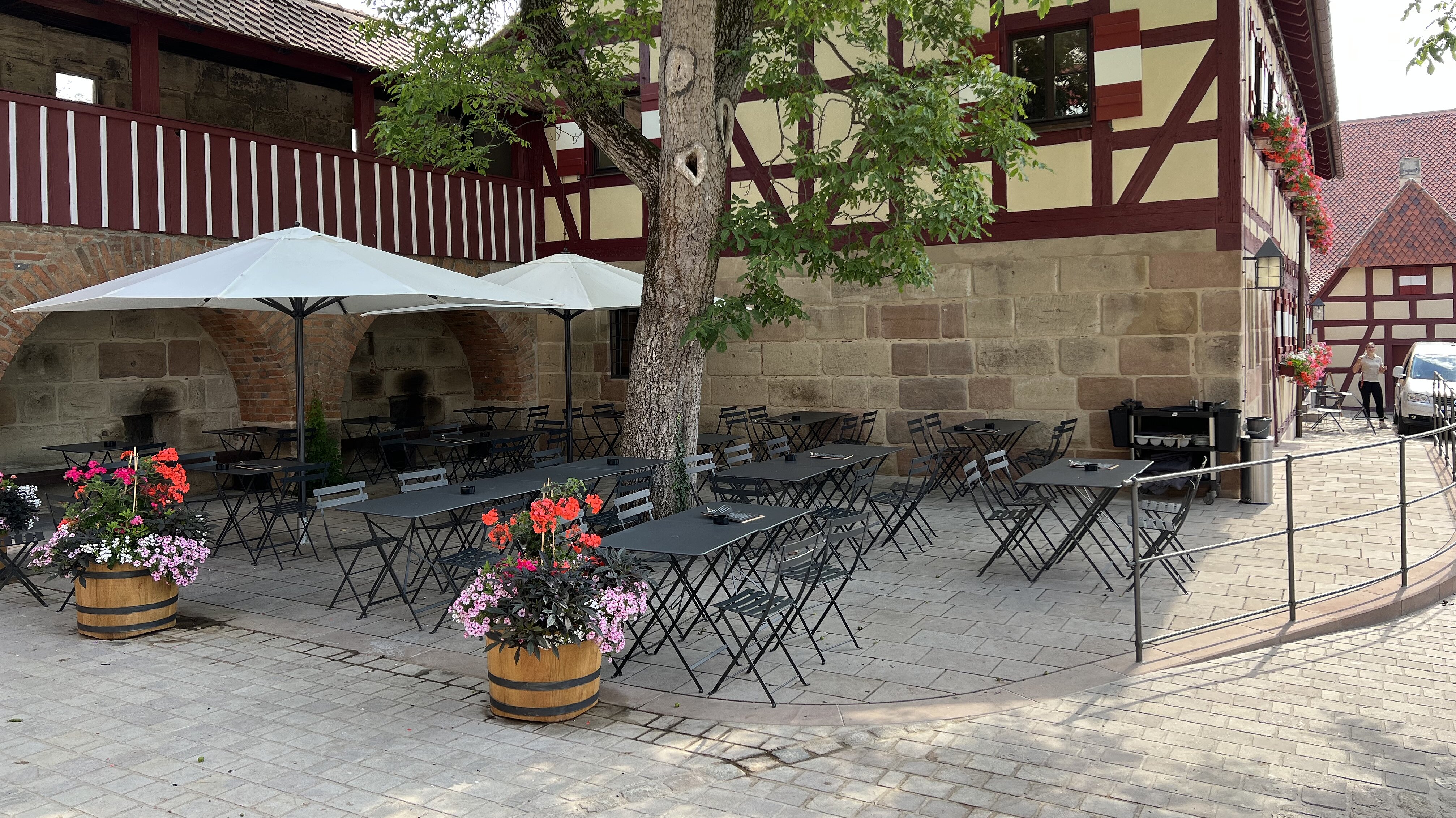 Der gepflasterte Außenbereich des Burgcafés Beer auf der Nürnberger Kaiserburg bietet schattige Sitzplätze unter Bäumen vor einer historischen Fachwerkfassade.