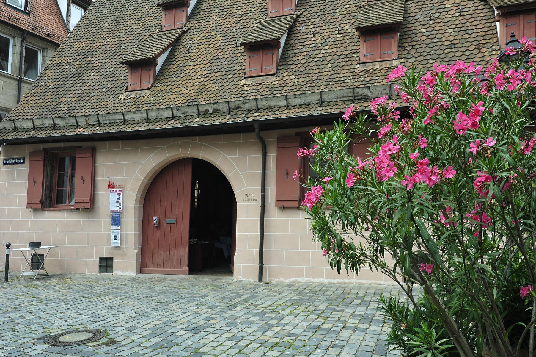 Das Gebäude der Kulturscheune der Altstadtfreunde hat ein rundbogiges Holztor, rötliche Klappläden und ein hohes Steildach, davor steht ein rosa blühender Oleanderstrauch.