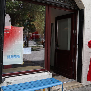 Offener Ladeneingang mit rotem Kleid auf Schaufensterpuppe rechts und blauem Holzbänkchen davor