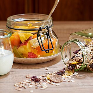 Drei Glasgefäße auf einem Holztisch zeigen Joghurt, bunten Fruchtsalat und Müsli, das teilweise aus einem umgekippten Glas herausgefallen ist.