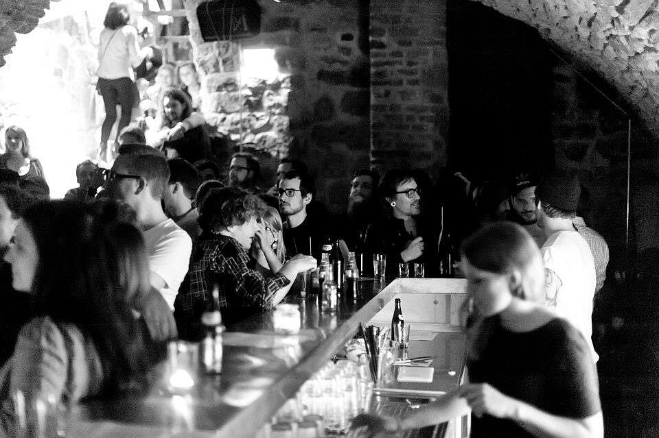 A black and white photograph of a busy bar inside a historic vaulted cellar, with guests standing at the counter and talking.