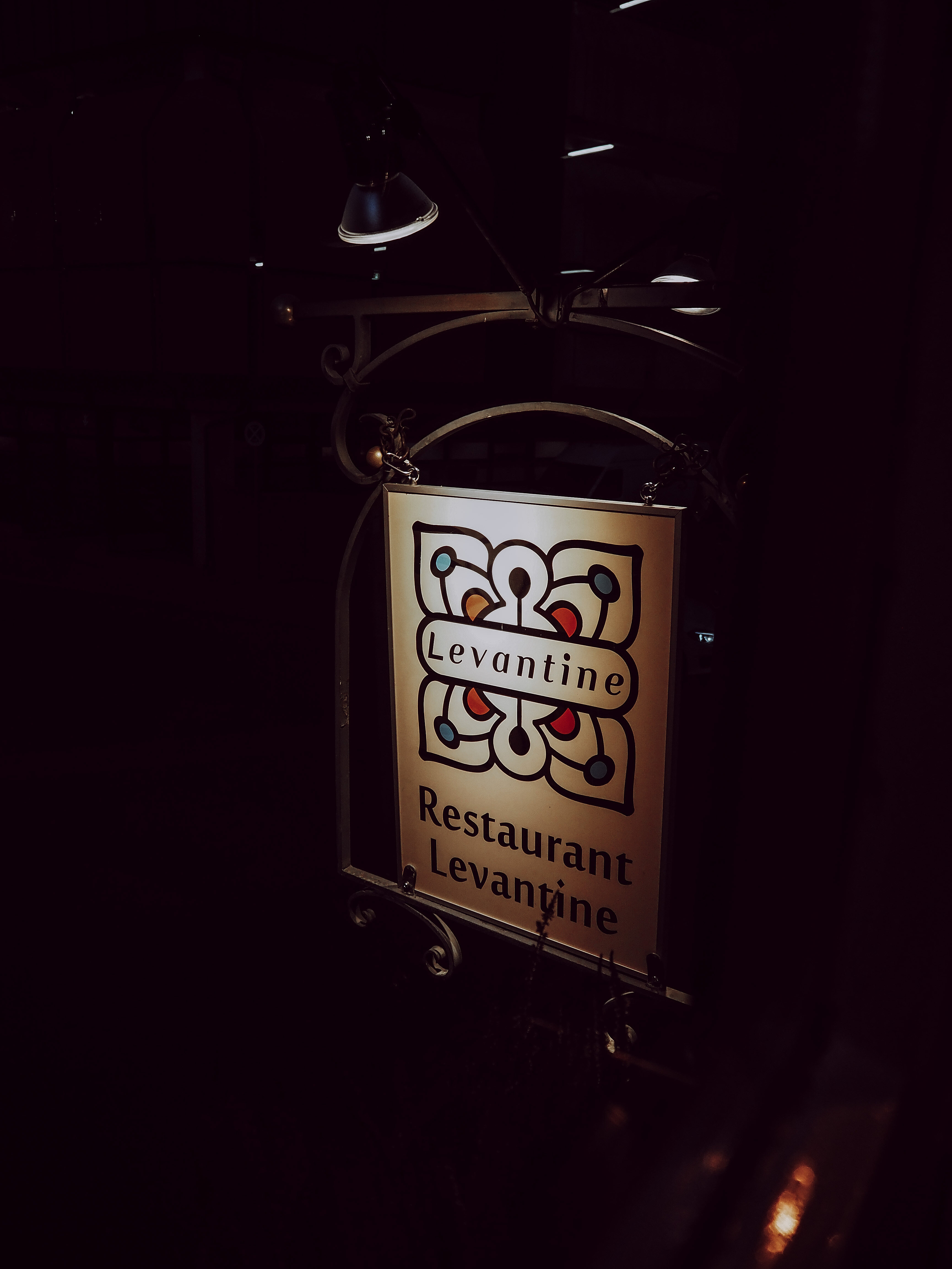 Ein beleuchtetes Schild des Restaurant Levantine hängt an einer schmiedeeisernen Halterung vor dunklem Hintergrund. Das Schild zeigt das farbige Logo des Levantine.