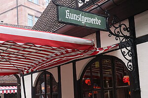 Außenansicht der Kerzenwerkstatt im Nürnberger Handwerkerhof mit einem grünen Schild „Kunstgewerbe“ an der Fachwerkfassade.