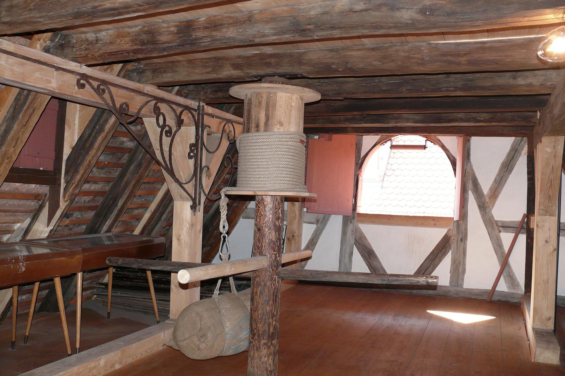 Historischer Dachboden der Kulturscheune der Altstadtfreunde mit massiven Deckenbalken, einer hölzernen Seilwinde im Zentrum und einem Blick durch ein Rundbogenfenster auf benachbarte Ziegeldächer.