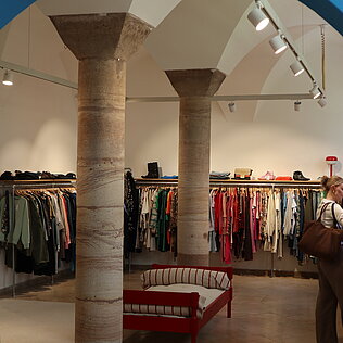 Blick durch einen blauen Torbogen auf einen Secondhand-Laden mit zwei steinernen Säulen, Kleiderstangen mit hängender Kleidung und einer roten Sitzbank in der Mitte