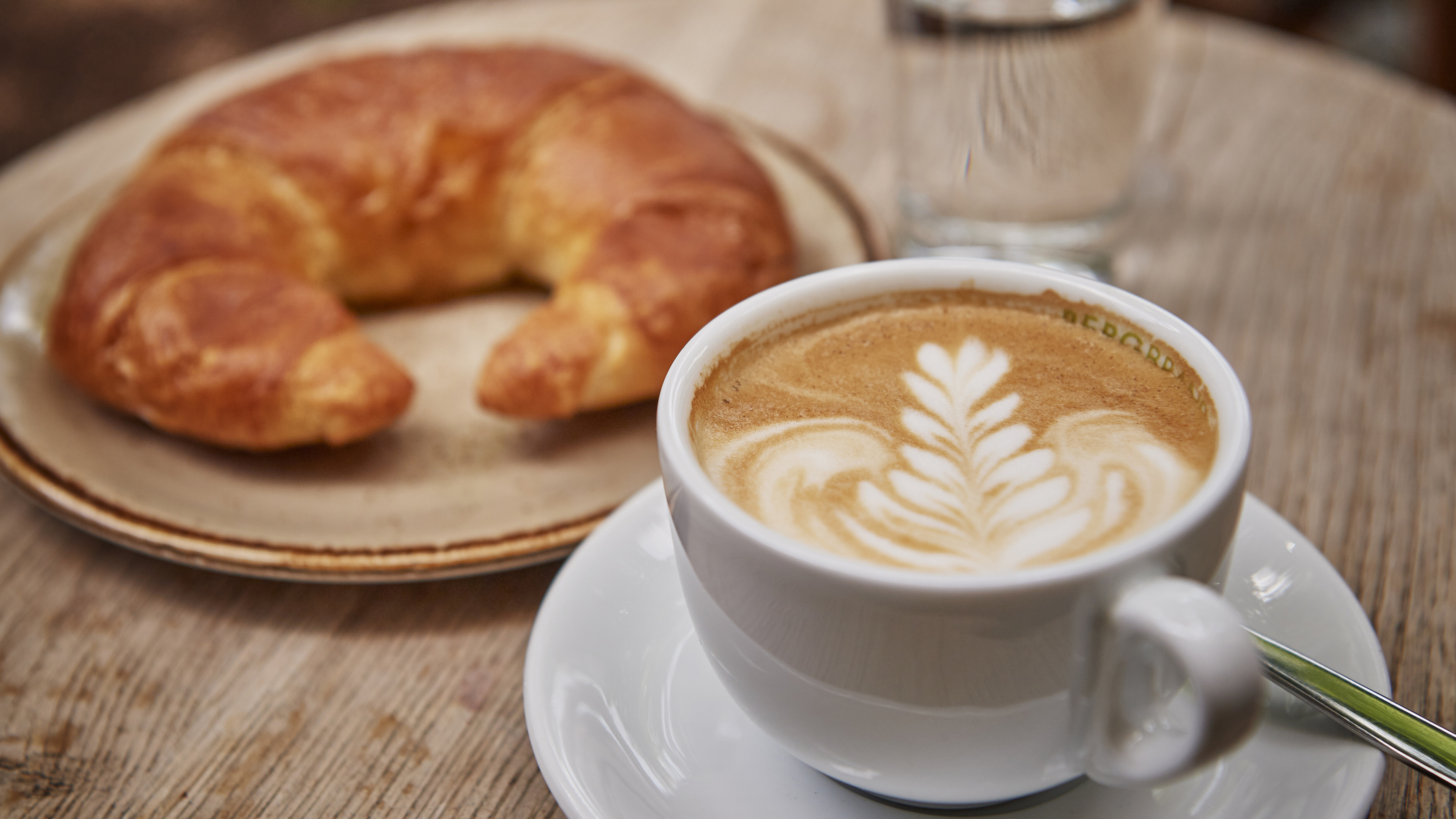 Ein Cappuccino mit Latte Art in einer weißen Tasse steht neben einem goldbraunen Croissant und einem Glas Wasser auf einem runden Holztisch.