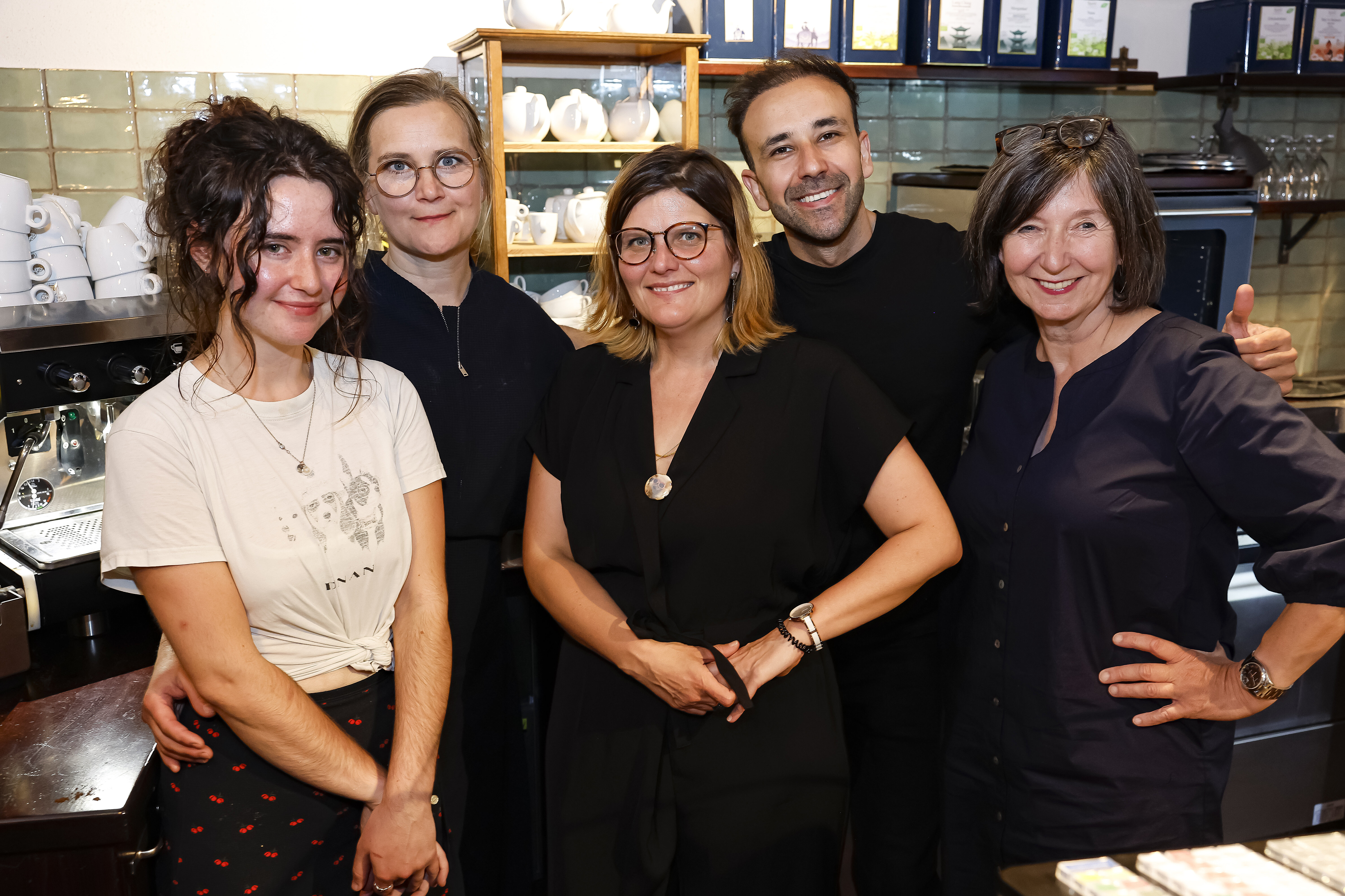 Das Team der Maulbeere, bestehend aus drei Frauen und einem Mann, lächelt gemeinsam für ein Gruppenfoto vor der Kaffeemaschine und dem Teeregal im Gastraum.