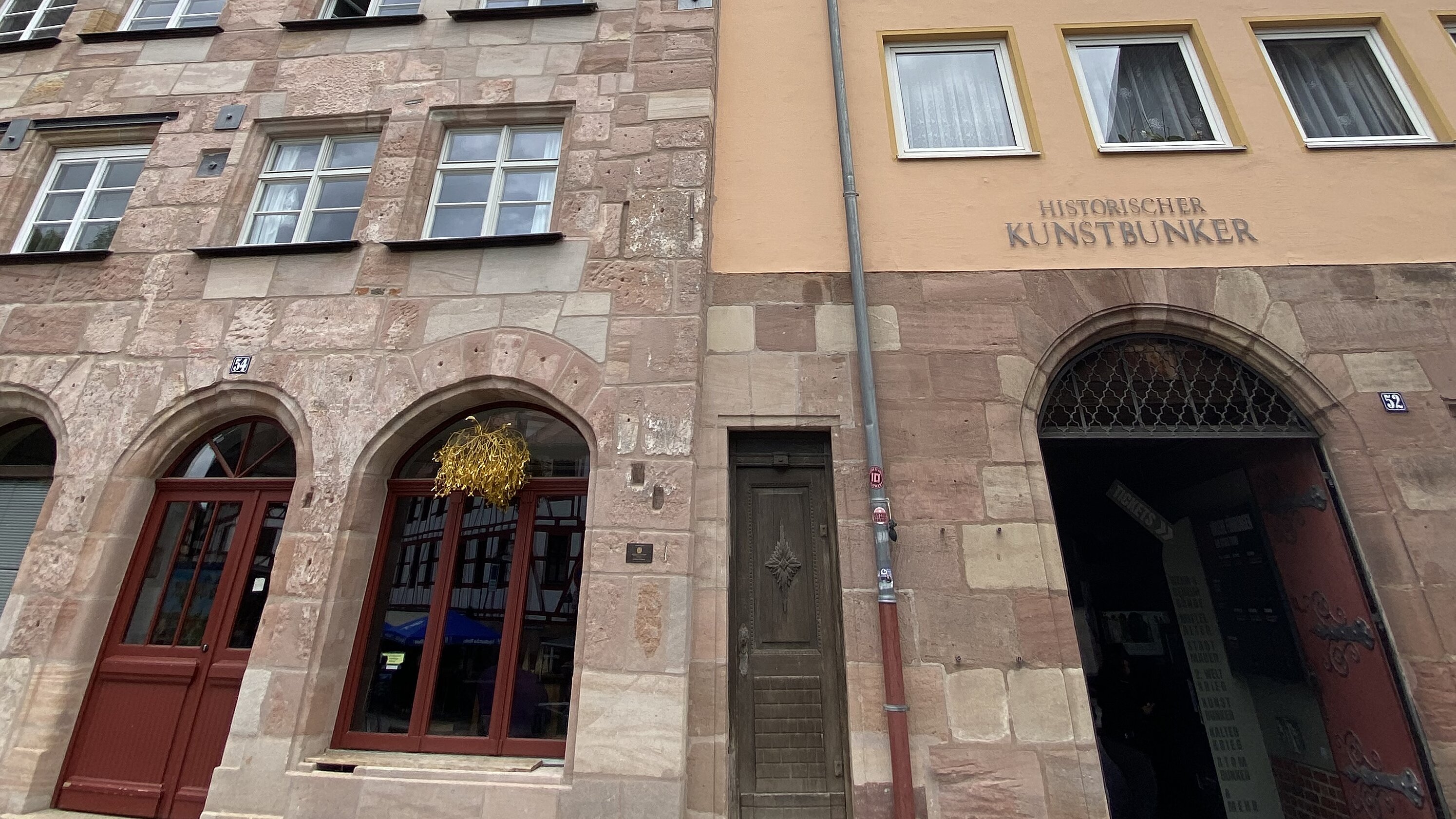 Der Eingang zum Historischen Kunstbunker liegt in einem Sandsteingebäude mit Rundbogenportal neben einem schmalen Altbau mit spitzem Giebel.