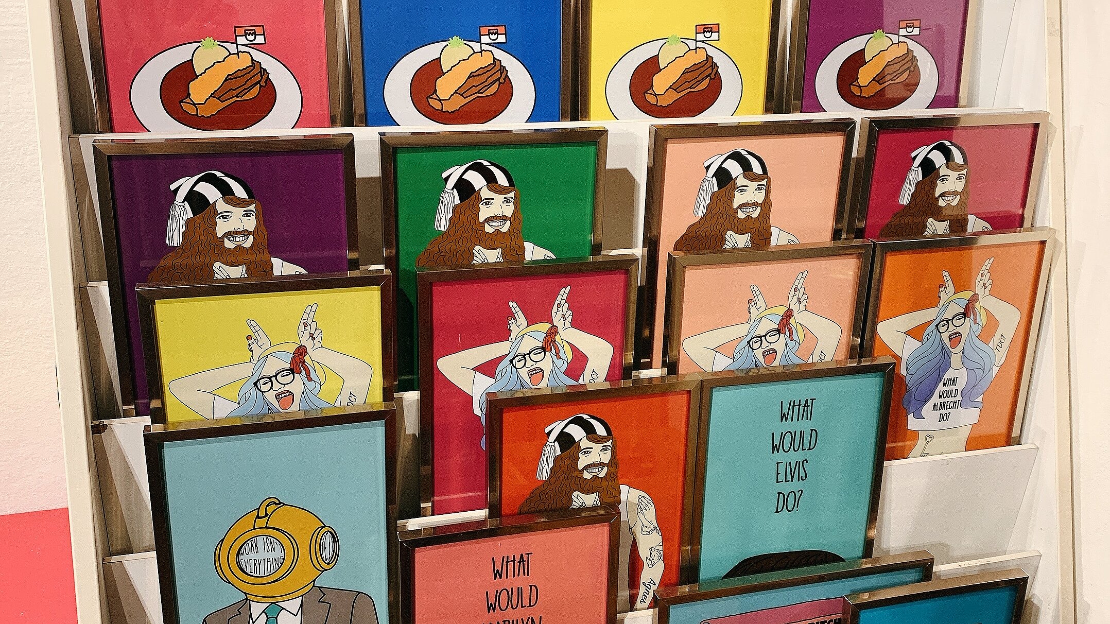 Ein weißes Verkaufsregal in der Bogi Nagy Galerie & Atelier ist mit zahlreichen gerahmten, farbenfrohen Kunstdrucken und Illustrationen im Pop-Art-Stil bestückt.
