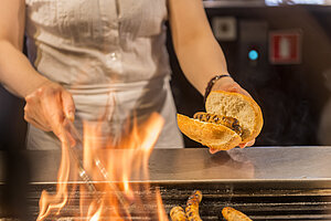 Nahaufnahme der Zubereitung von Nürnberger Rostbratwürsten über offenem Feuer: Eine Person hält eine frisch gegrillte Bratwurst in einem aufgeschnittenen Brötchen über lodernden Flammen.