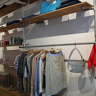 Kleidung auf Holzregalen und Kleiderstangen in einem Secondhand-Laden mit einer roten Jacke und einer grauen Tasche an der Wand