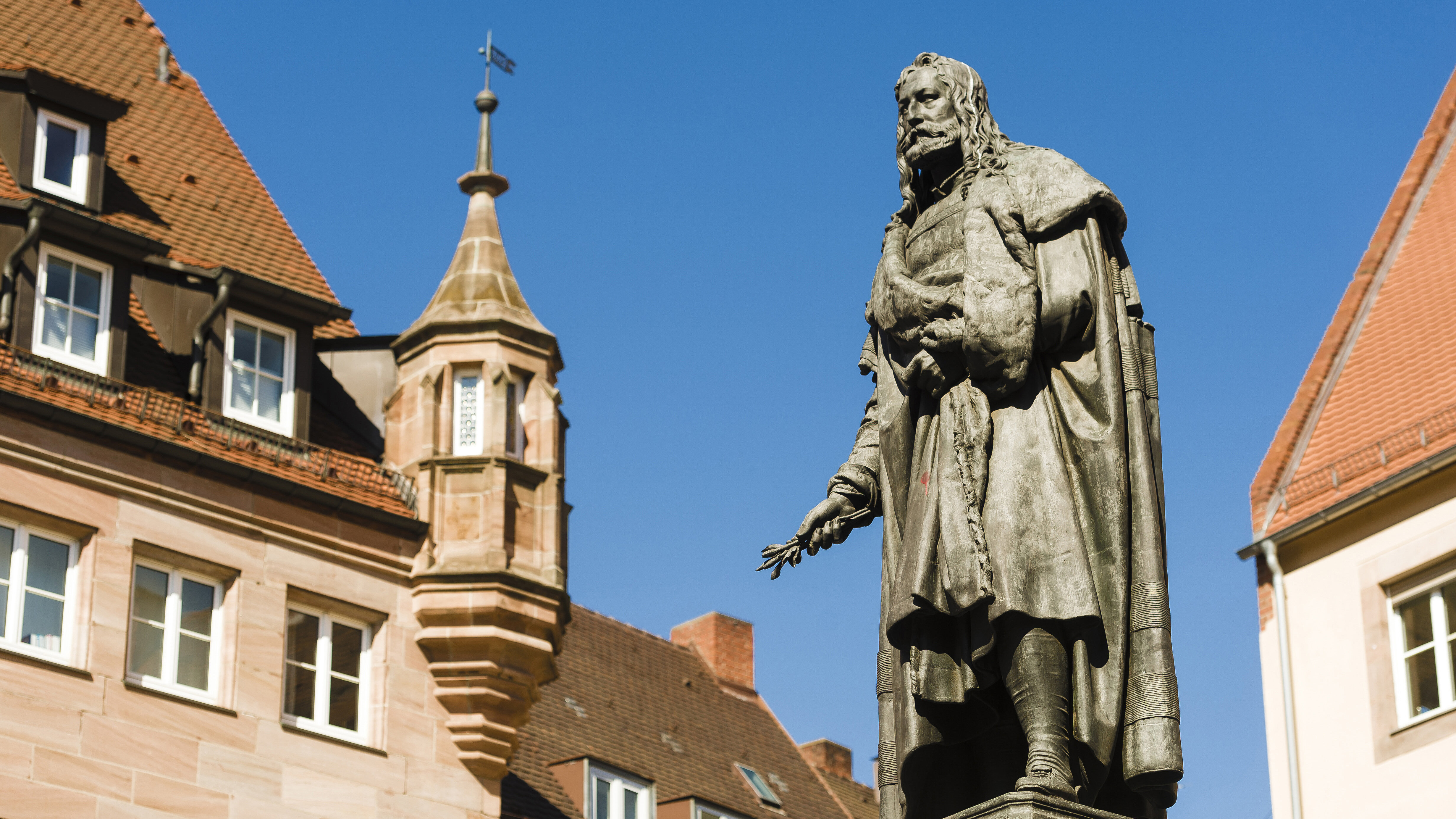 Bronze-Standbild von Albrecht Dürer auf einem steinernen Sockel vor dem Hintergrund historischer Sandsteingebäude unter einem klaren blauen Himmel.
