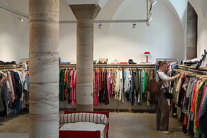 Innenansicht eines Secondhand-Kleidungsgeschäfts mit mehreren Kleiderstangen, zwei Frauen beim Stöbern und zwei großen Säulen im Raum