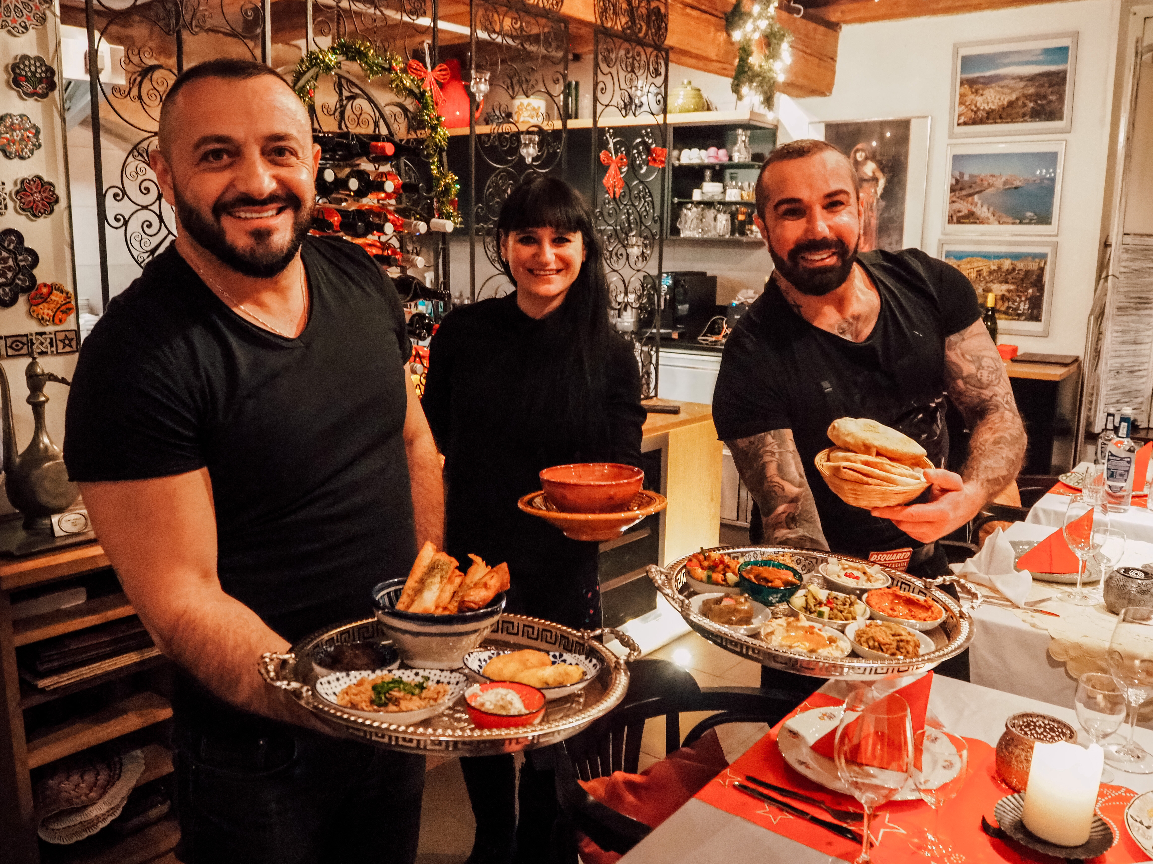 Drei lächelnde Mitarbeiter des Levantine stehen im Gastraum und präsentieren eine Auswahl an Speisen auf verzierten silbernen Tabletts sowie in Schälchen und Brotkörben.
