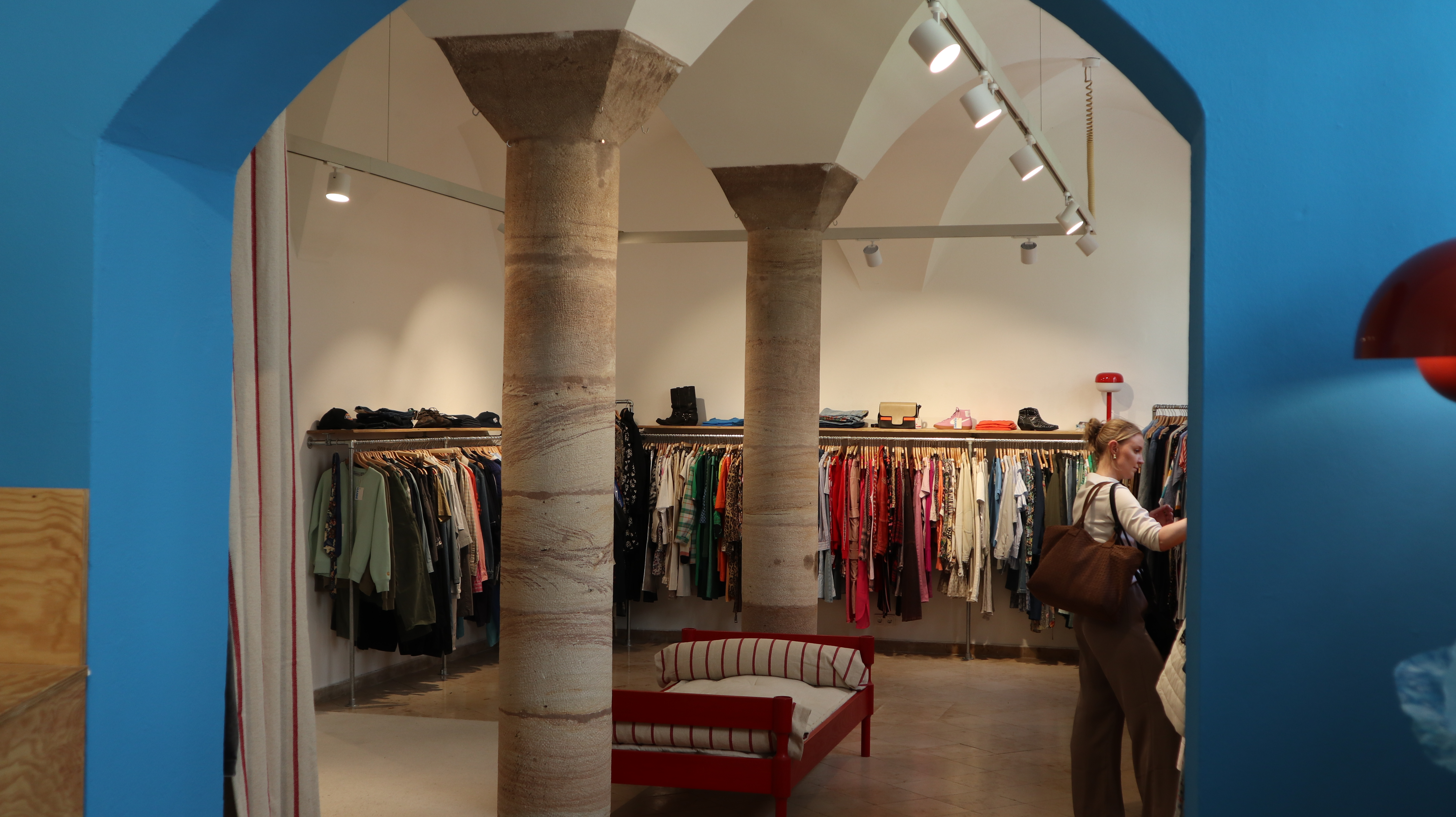 Blick durch einen blauen Torbogen auf einen Secondhand-Laden mit zwei steinernen Säulen, Kleiderstangen mit hängender Kleidung und einer roten Sitzbank in der Mitte