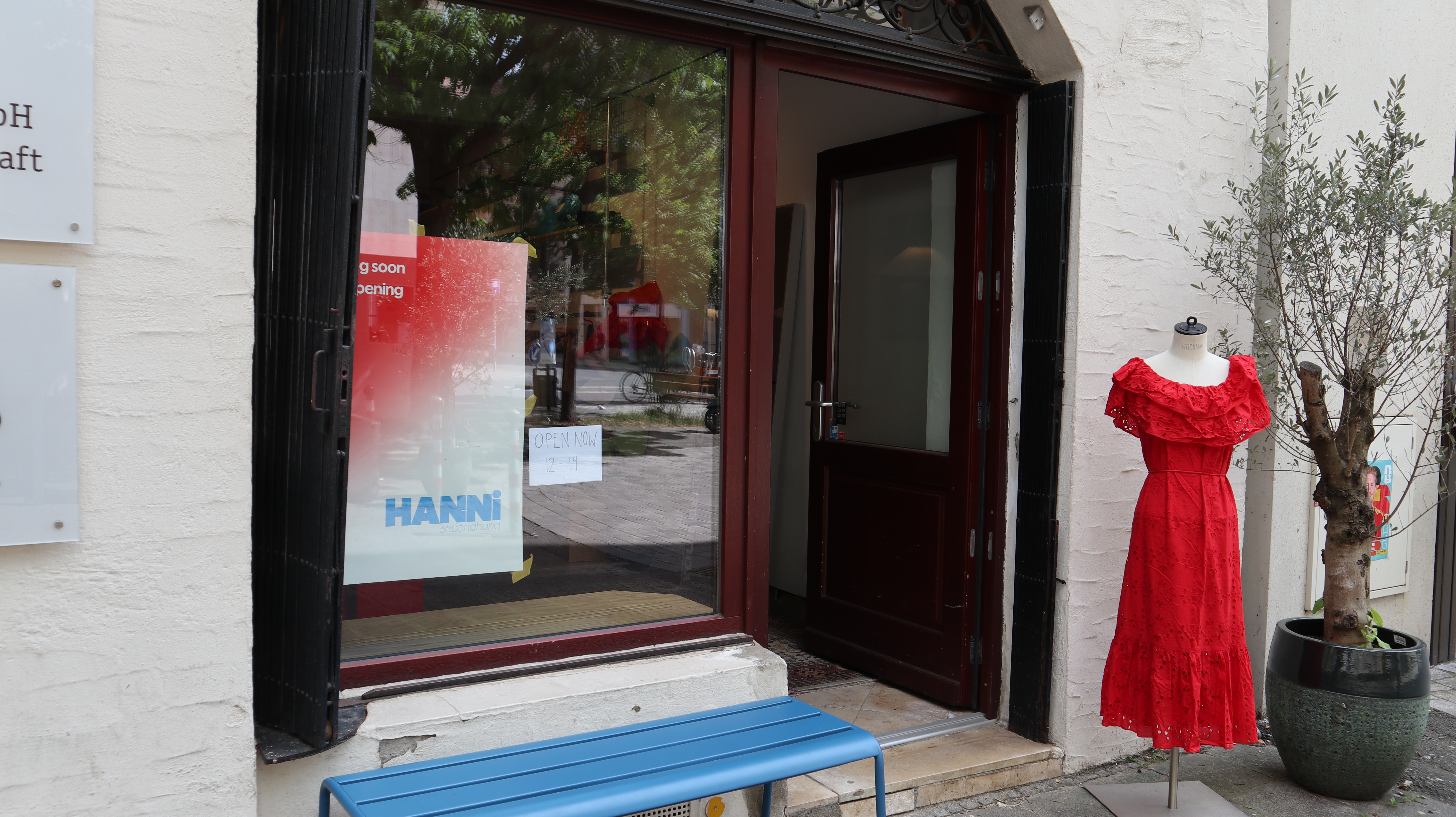 Offener Ladeneingang mit rotem Kleid auf Schaufensterpuppe rechts und blauem Holzbänkchen davor