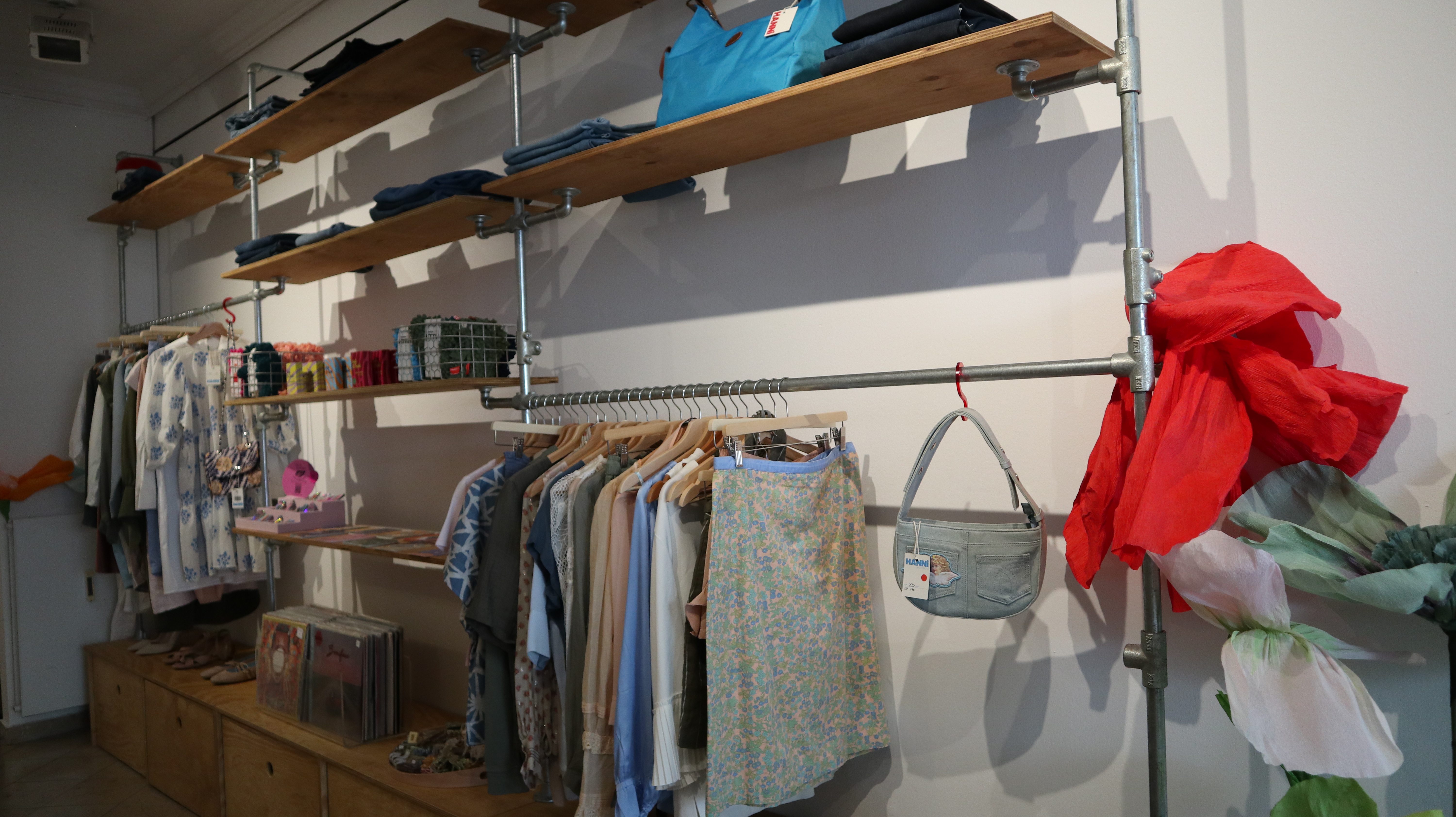 Kleidung auf Holzregalen und Kleiderstangen in einem Secondhand-Laden mit einer roten Jacke und einer grauen Tasche an der Wand