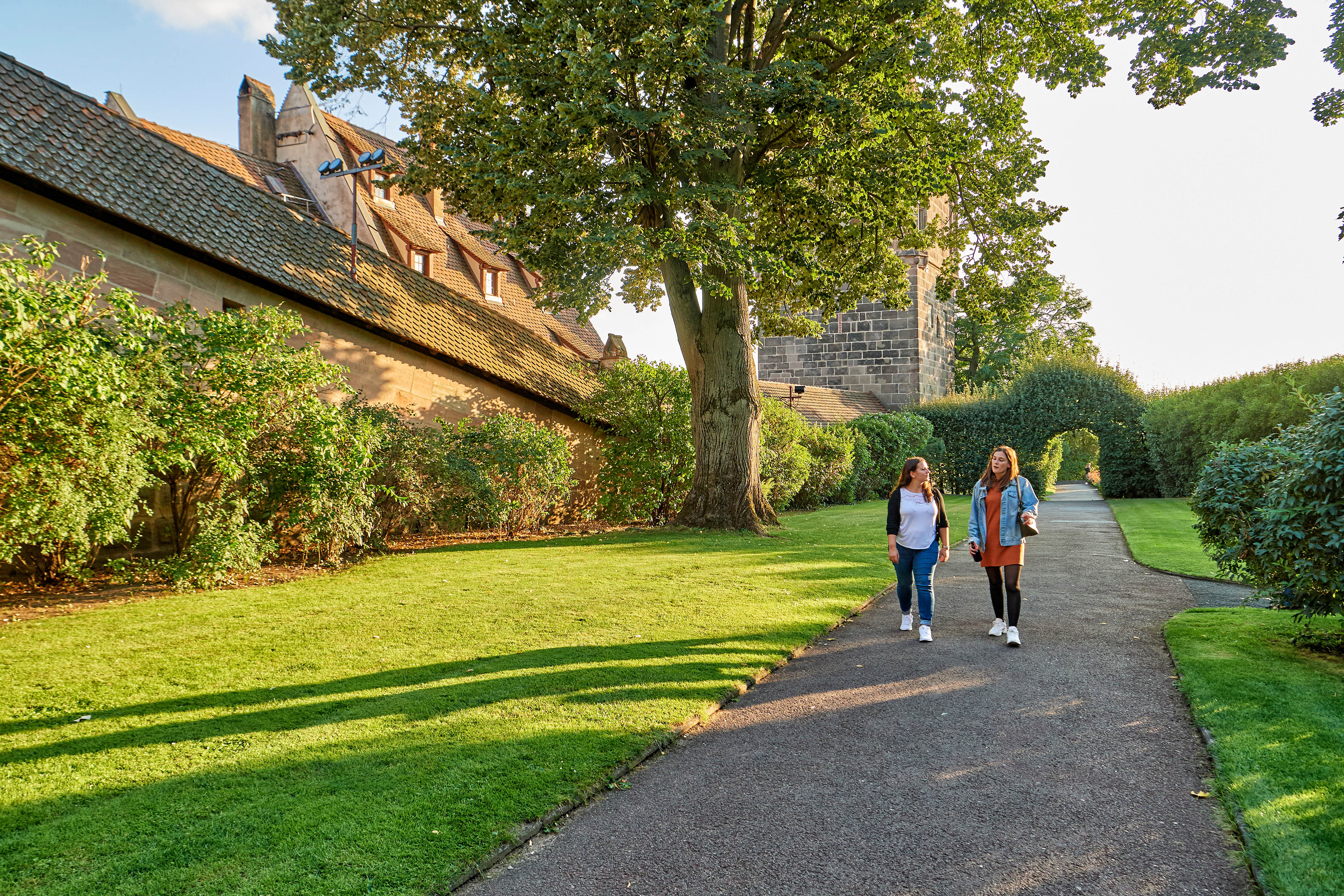 Zwei Frauen gehen auf einem Weg im Burggarten Nürnberg, umgeben von Bäumen, Hecken und historischen Mauern.